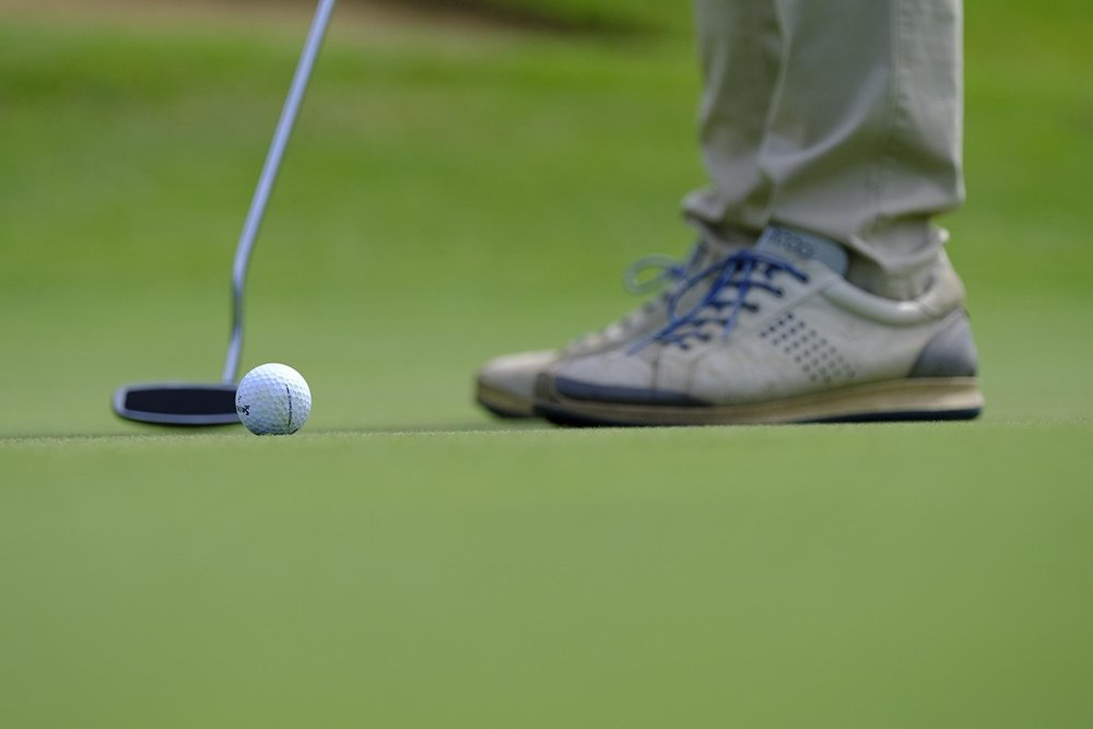 Putter set behind a golf ball on a green, golfer's shoes in view