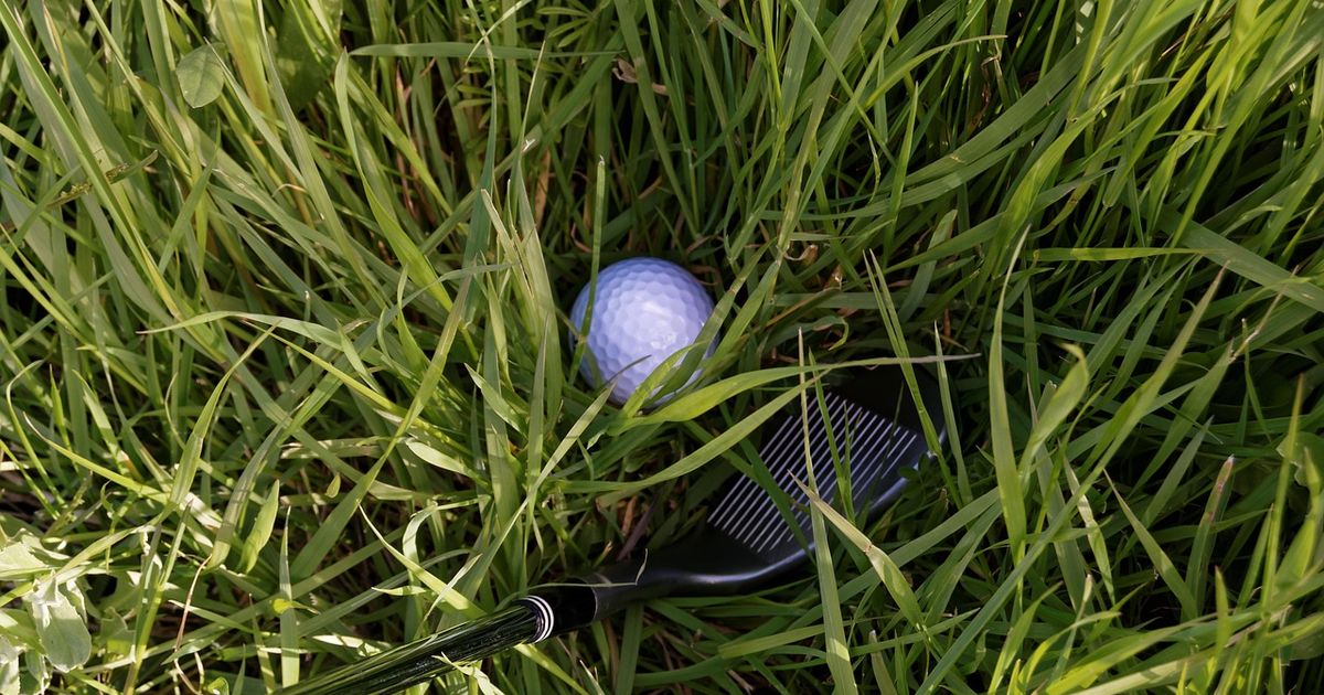 White golf ball sitting down in long grass next to a wedge