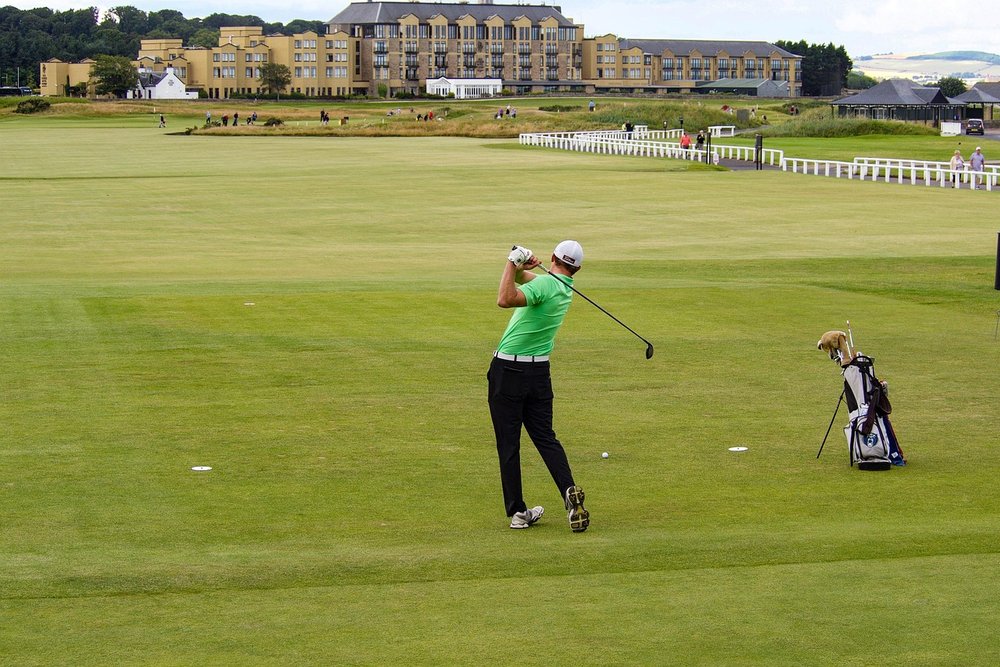 Golfer hitting a tee shot on a wide links fairway with a hotel behind
