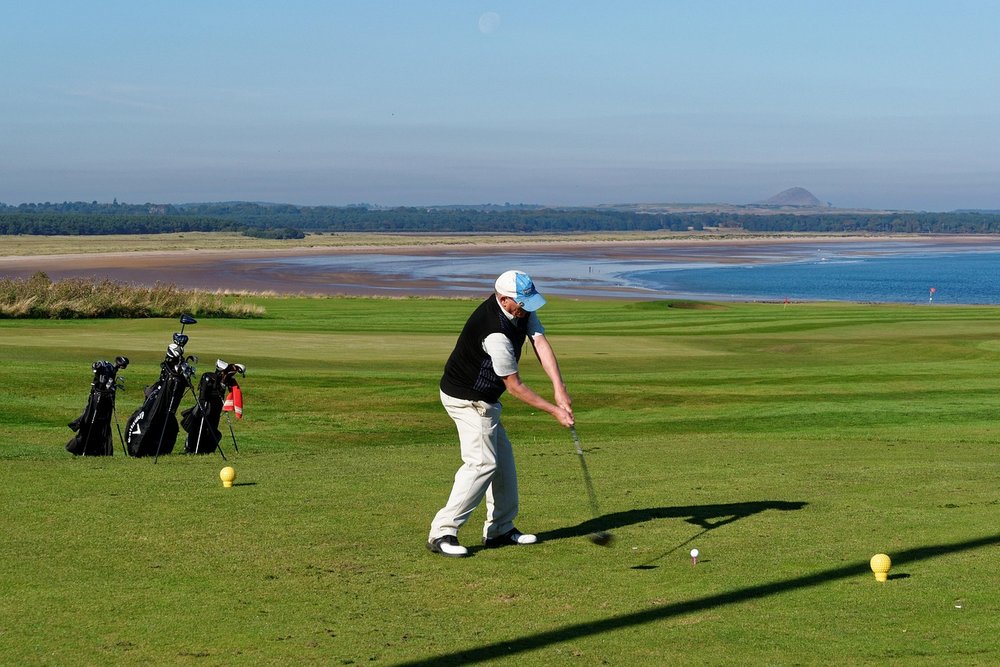 Golfer hitting a tee shot from the teeing area on a coastal course, with golf bags behind